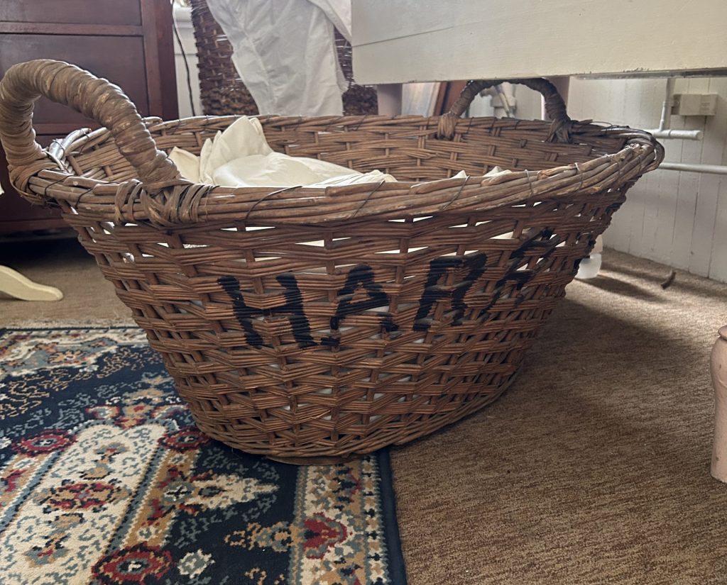 A large wicker basket with the name HARRIS stamped in black letters on the side. It is sitting on a blue floral rug.