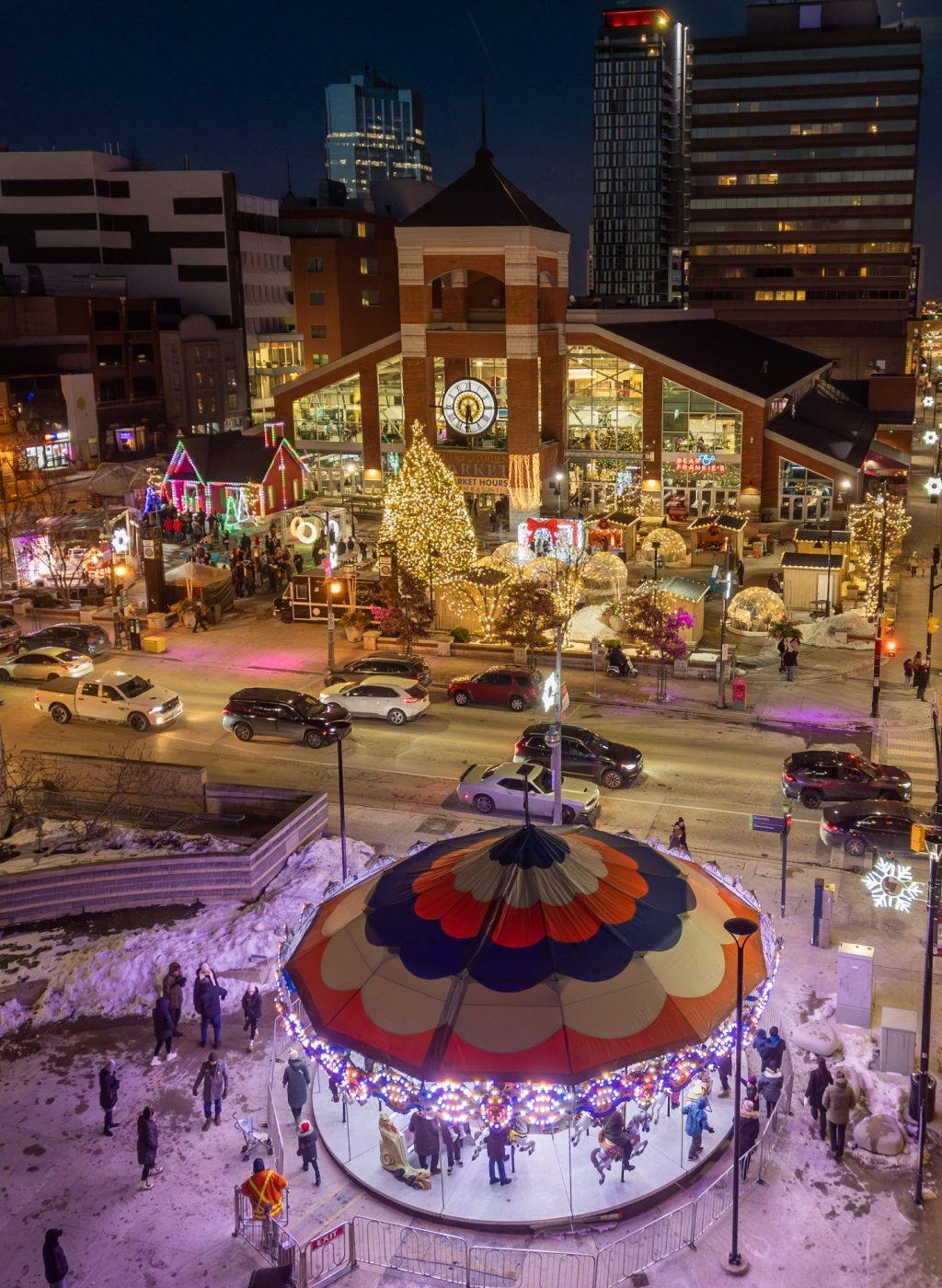 An image of a winter scene with a carousel in front and a large well lit red building behind. In front there are stalls and booths.