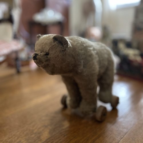 An image of a beige stuffed bear. He has wheels attached to his feet. He is sitting on a wooden floor, the background is blurred.