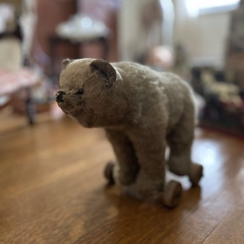 An image of a beige stuffed bear. He has wheels attached to his feet. He is sitting on a wooden floor, the background is blurred.