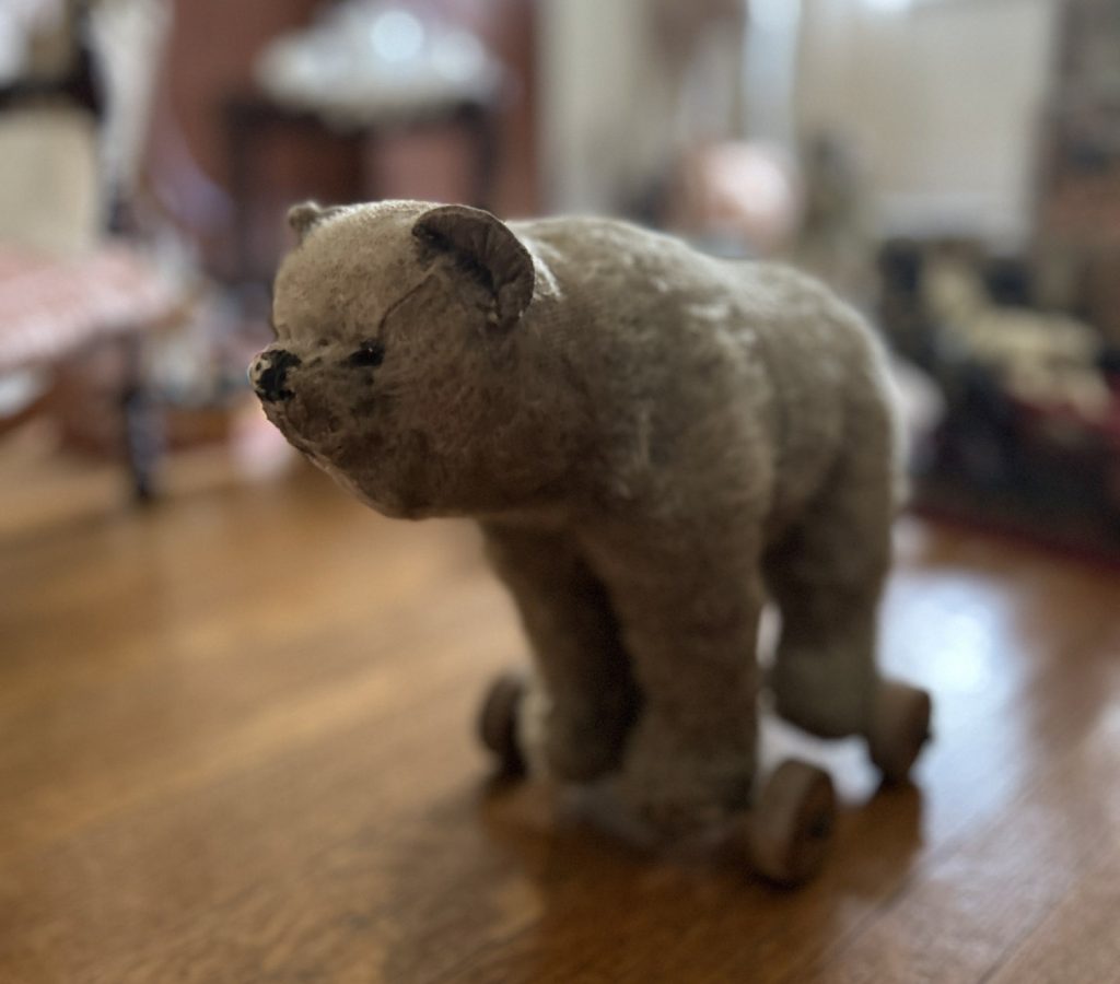 An image of a beige stuffed bear. He has wheels attached to his feet. He is sitting on a wooden floor, the background is blurred.