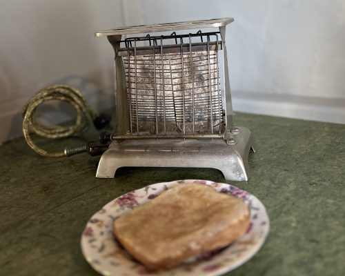 A metal wire toaster sitting on a green counter. In front is a floral plate with a slice of toast.